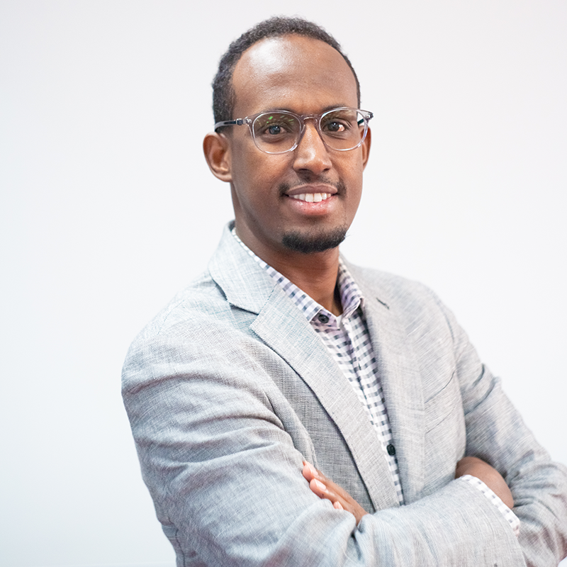 Mukhtar Ibrahim headshot, Mukhtar is standing in a gray suit jacket with his arms crossed.