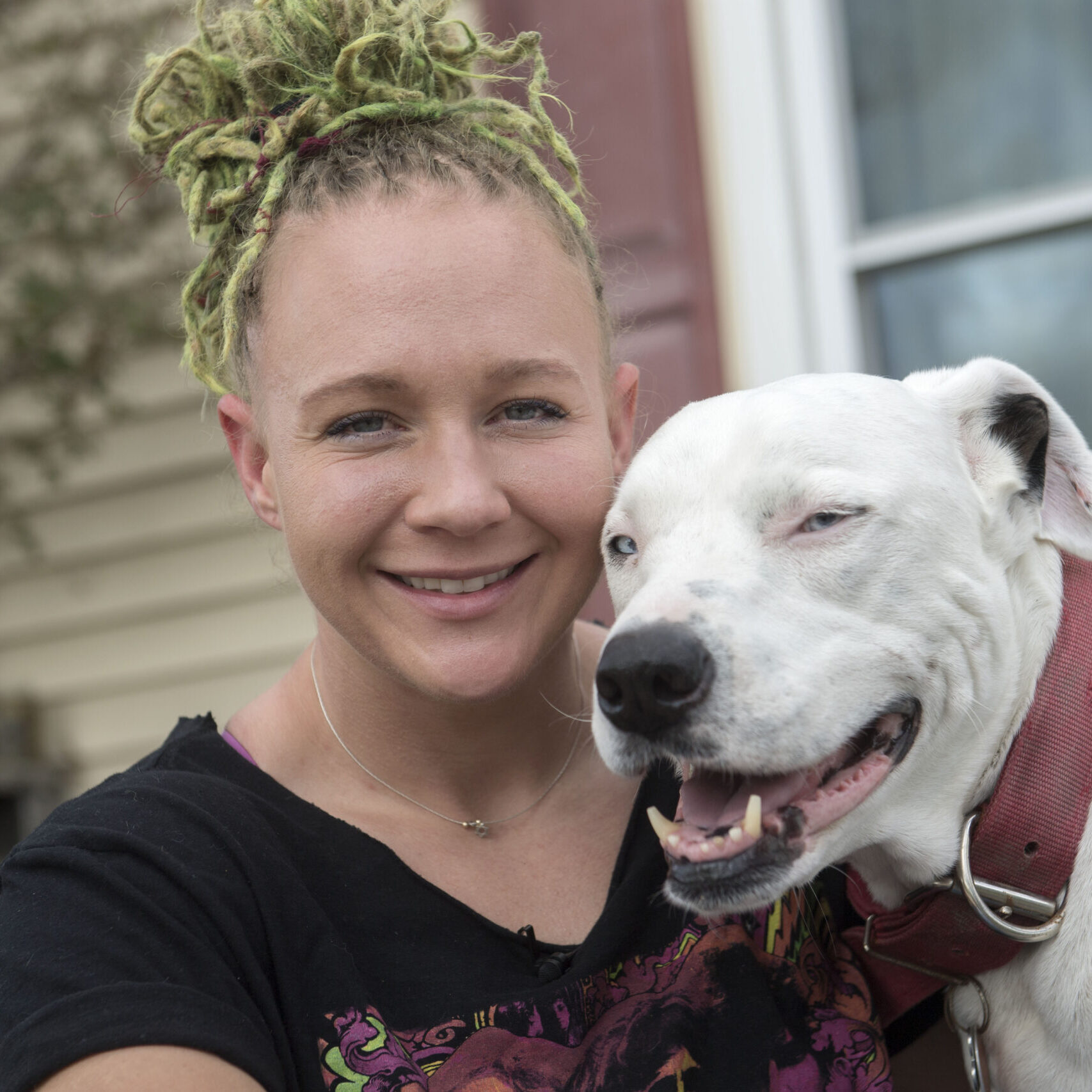 Reality winner sits on the porch, she is a white woman with dyed green braids, hugging a white dog. They're both smiling.