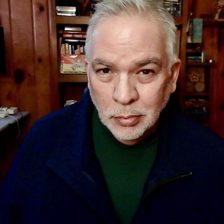 Ricardo Sandoval-Palos 2024 headshot (1) Close up headshot of Ricardo Sandoval-Palos with a bookshelf in the background.